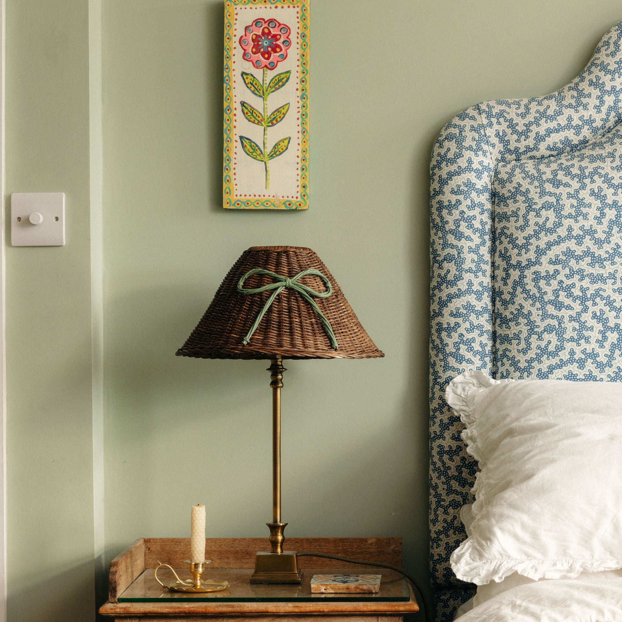 Wicker lamp on a wooden side table with floral wall art and patterned headboard.