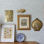 Decorative wall with framed artwork, gold fish and shell sculptures, and a blue and white plate against a patterned wallpaper.
