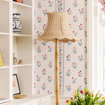 Cecelia Wicker floor lamp against a floral wallpapered wall with a shelf and flowers in the foreground.