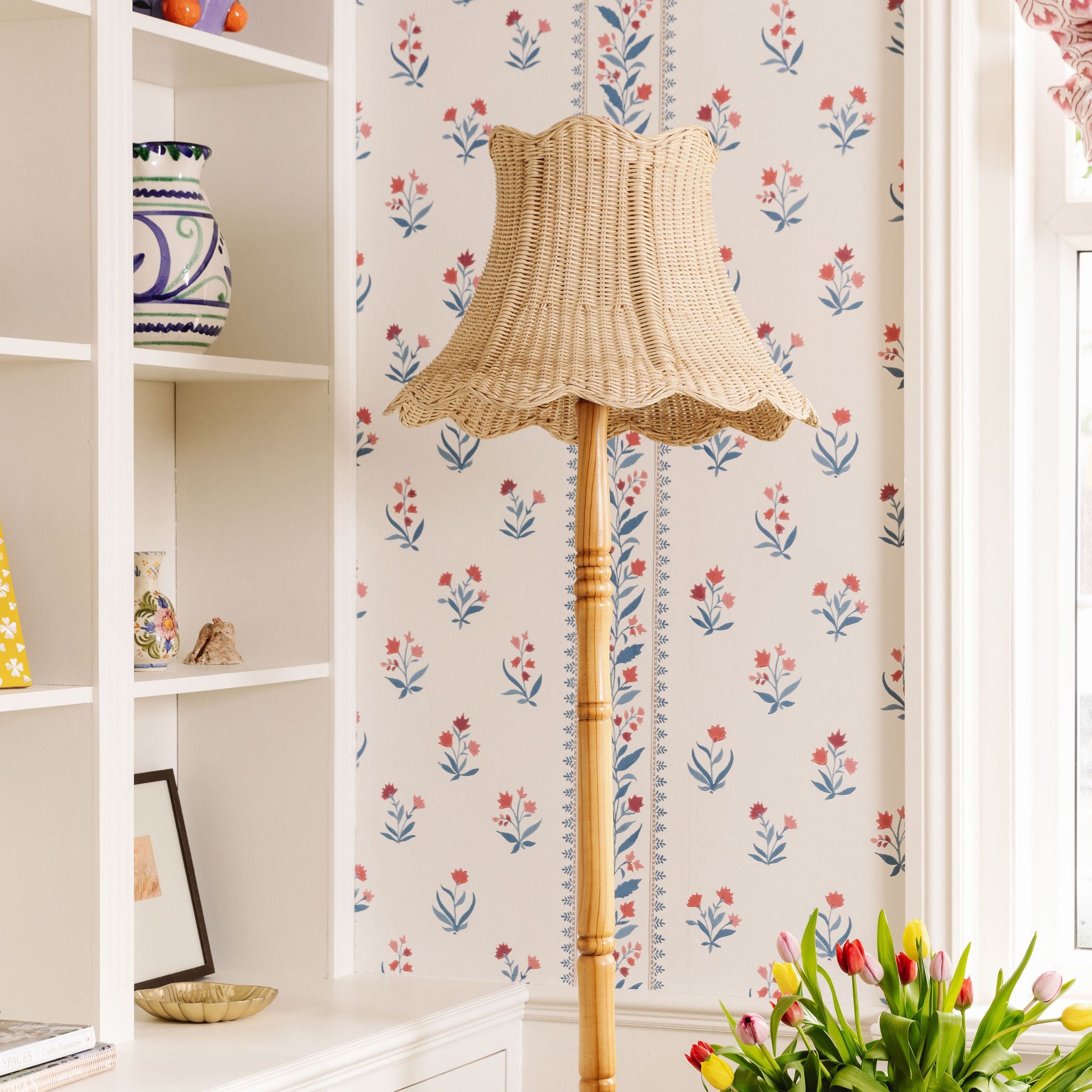 Cecelia Wicker floor lamp against a floral wallpapered wall with a shelf and flowers in the foreground.