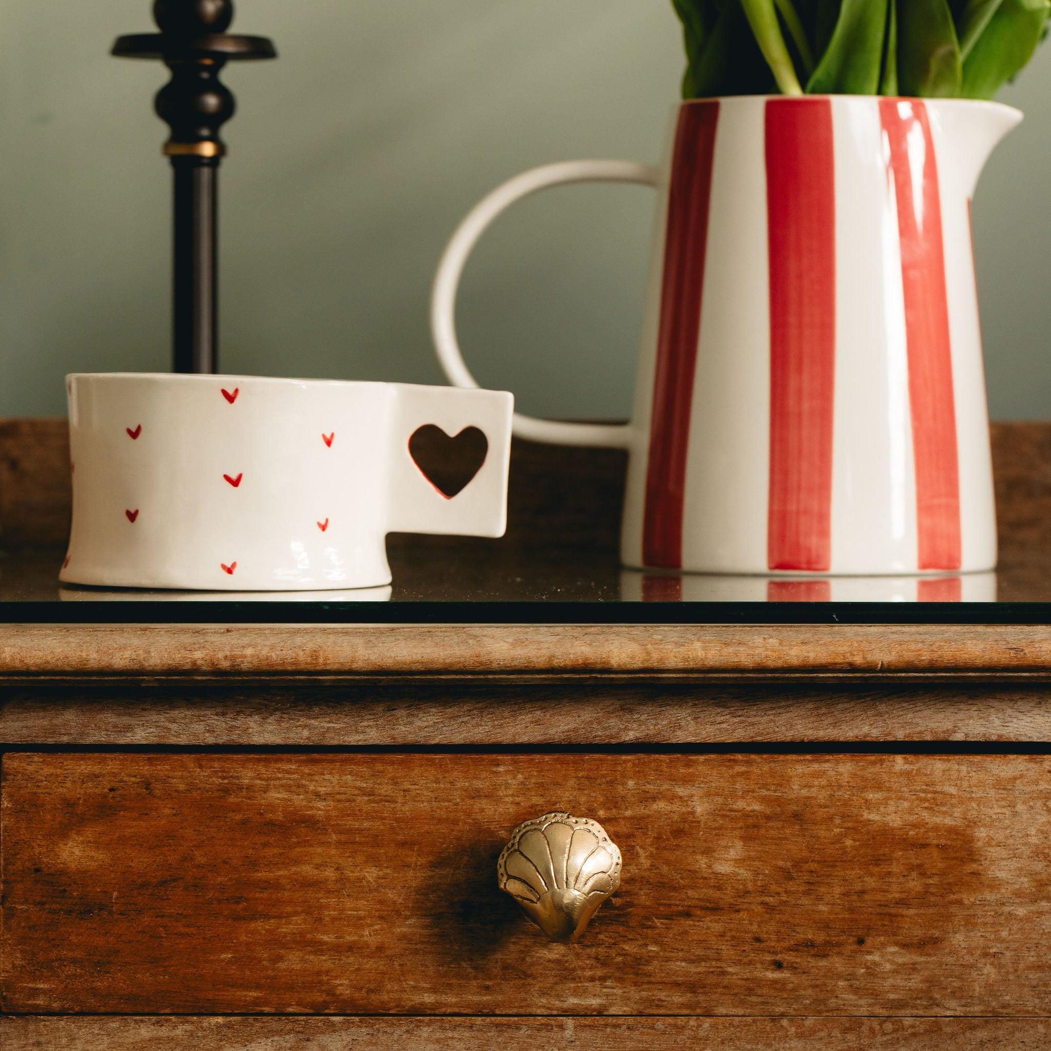 White mug with red hearts and a heart-shaped object on a wooden surface next to a striped pitcher.