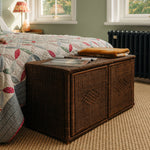 Wicker storage chest in a bedroom with a quilted bedspread and lamp.