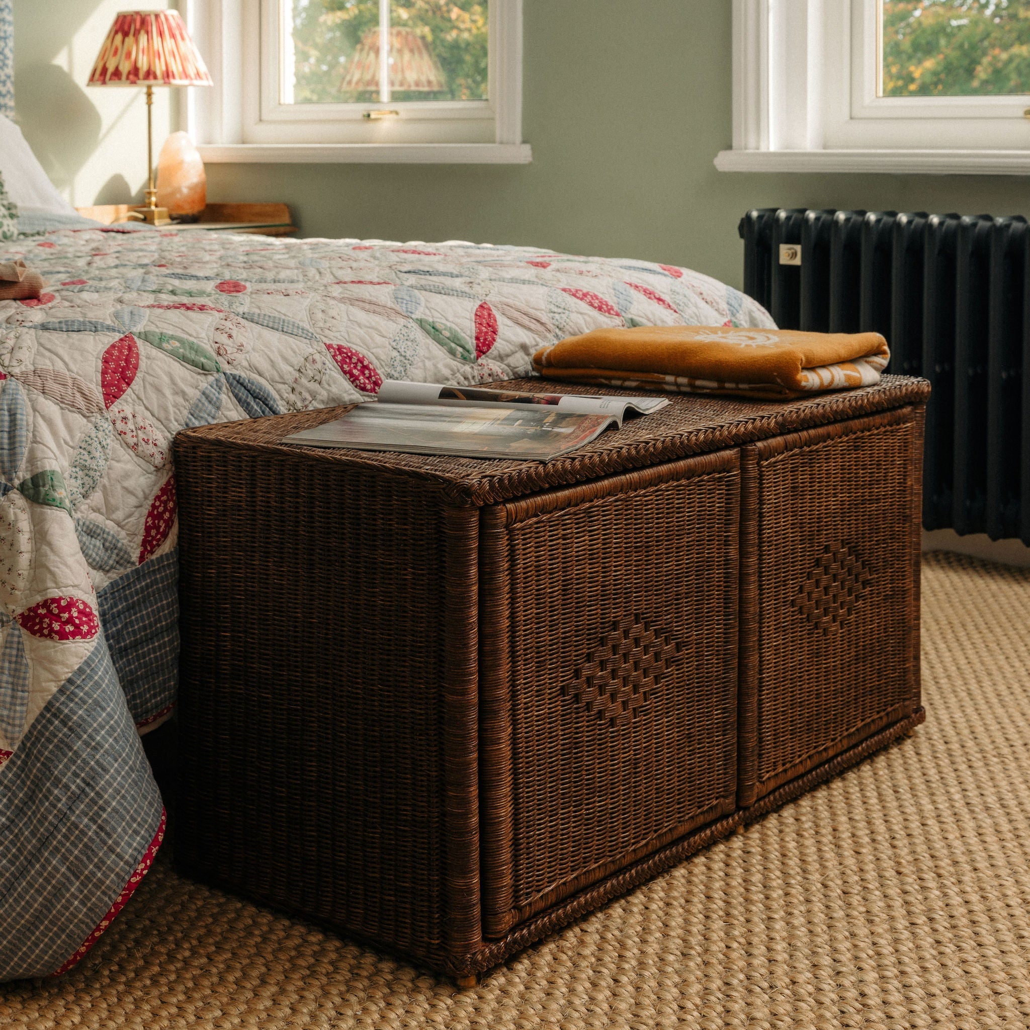 Wicker storage chest in a bedroom with a quilted bedspread and lamp.