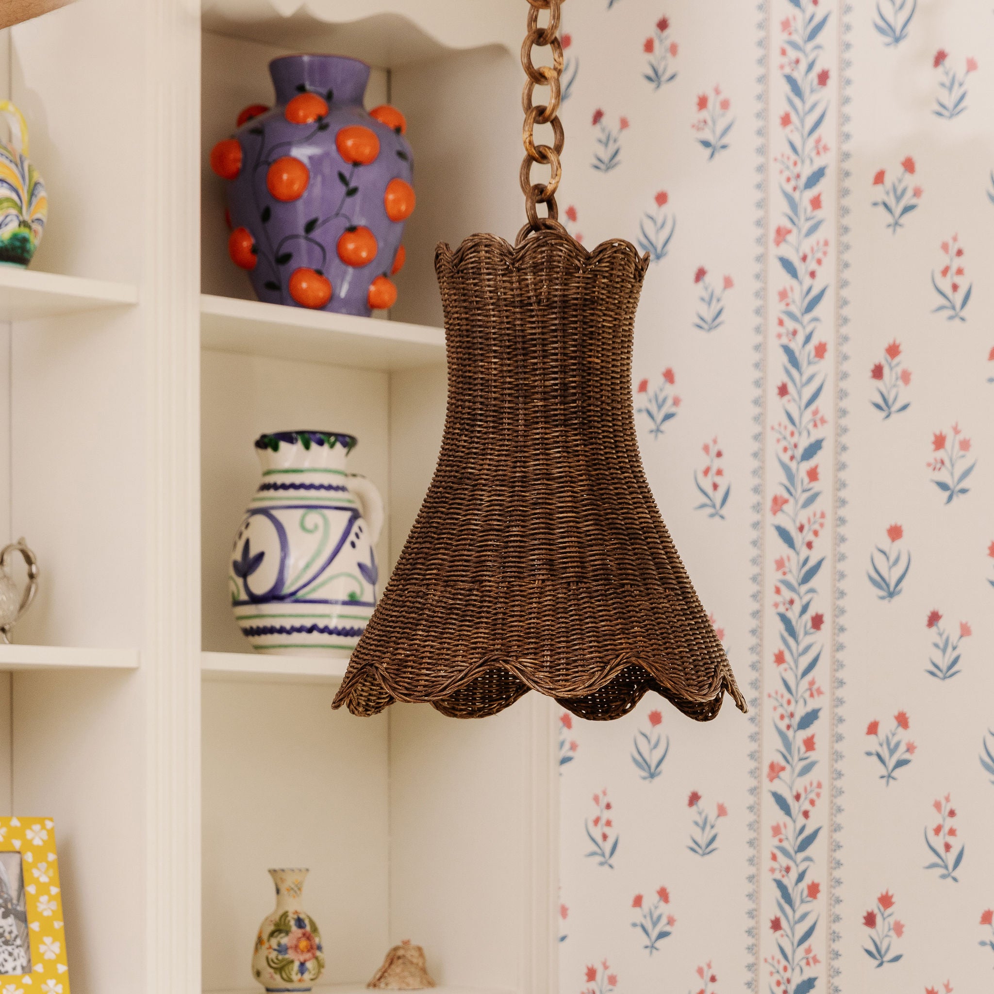 Calla Wicker pendant hanging in front of a shelf with decorative vases against a floral wallpapered wall.